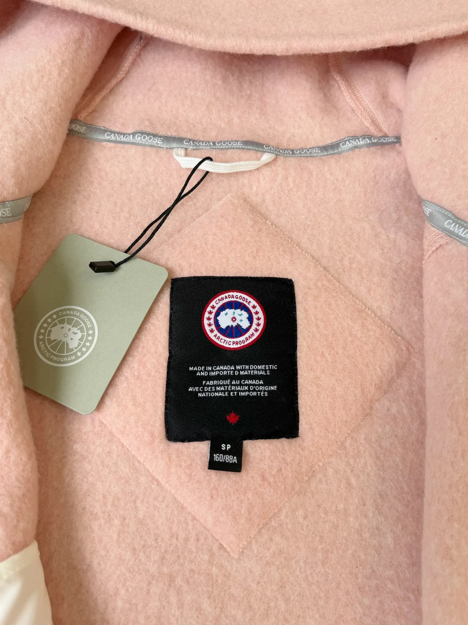 P660 Canada Goose Fleece Wool Badge Hooded Jacket Fabric selection: Wool fleece fabric blended with wool as the main body and a very small amount of spandex and polyester fiber. Wool is a core natural ingredient that provides unparalleled temperature regulation, antibacterial and anti-odor properties and a skin-friendly feel. The feeding of a small amount of spandex gives the fabric a certain degree of four-way elasticity, making it non-restrictive during daily activities. As the basis of fleece's structure, polyester fiber enhances the fabric's durability, wrinkle resistance and quick-drying properties. It contributes to the loft of the fleece and provides functions such as trapping air.  Detail 1: The sleeves and hem are closed with 2×2 ribbing. Its strong elasticity can closely fit wrists and waists of different thicknesses, forming an effective 'seal' to prevent cold wind from blowing back. It can still maintain its original shape after repeated stretching and use, and is not easy to loose and deform. And the ribbed cuffs can convey a sense of design and enhance the visual beauty.  Detail 2: The combination of hood design and elastic buckle improves warmth and windproof performance when wearing. Accessories: Customized elastic drawstring, elastic buckle, custom zipper pendant, custom armband Size: S M L Color: black gray coffee rice pink
